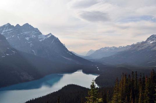 Nestled in the heart of the Canadian Rockies, this breathtaking vista captures the serene beauty of a glacier-fed lake surrounded by towering, snow-capped peaks. The lake, with its striking turquoise waters, is a tranquil gem set amidst the rugged wilderness. Its color, a result of glacial silt, stands in stark contrast to the dark, evergreen forests that blanket the surrounding slopes.  The mountains, with their jagged profiles, rise majestically, their summits etched against the soft hues of the sky. These peaks, carved over millennia by the relentless forces of nature, tell a silent story of geological grandeur and timelessness. The interplay of light and shadow across the landscape highlights the texture and contours of the rocky slopes, adding depth and dimension to the scene.  A thin layer of mist hangs over the distant mountains, creating a dreamy, ethereal atmosphere. This mist, combined with the gentle ripples on the lake's surface, suggests a moment of calm and stillness, inviting viewers to pause and soak in the natural splendor. The clarity of the water reflects the surrounding scenery like a mirror, enhancing the sense of purity and unspoiled beauty.